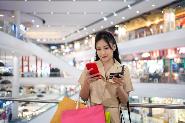 Фото Молодая женщина в умной повседневной одежде, несущая много бумажных пакетов для покупок, покупает онлайн на мобильном телефоне, используя кредитную карту в торговом центре