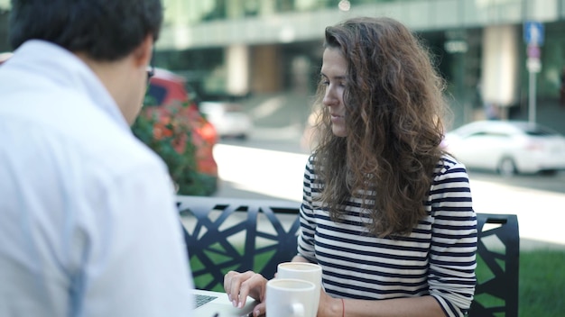 Фото Молодая женщина в полосатой рубашке и ее красивый коллега в белой рубашке работают вместе в летнем парке с ноутбуком и буфером обмена мужчина пьет кофе