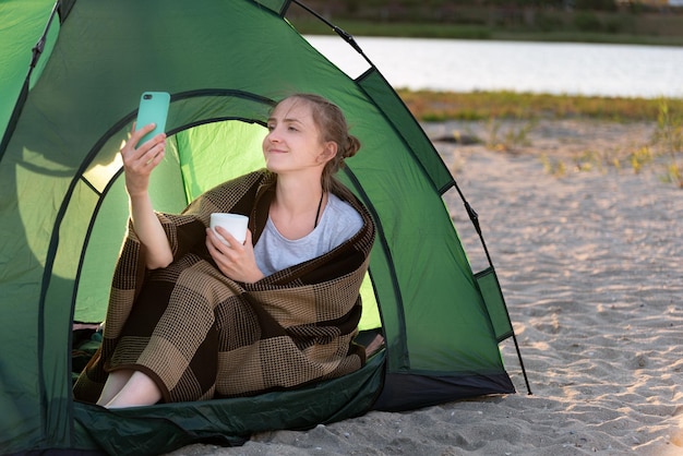 사진 텐트에서 커피를 마시고 셀카를 찍는 젊은 여성. 모래 해변에서 캠핑.