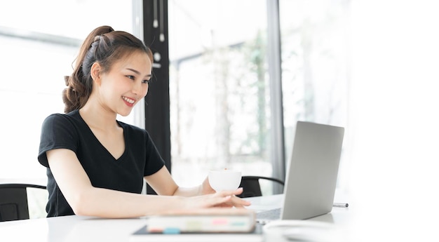 写真 シャツを着た若い女性がラップトップコンピュータを使いオフィスでコーヒーを飲んで座っています