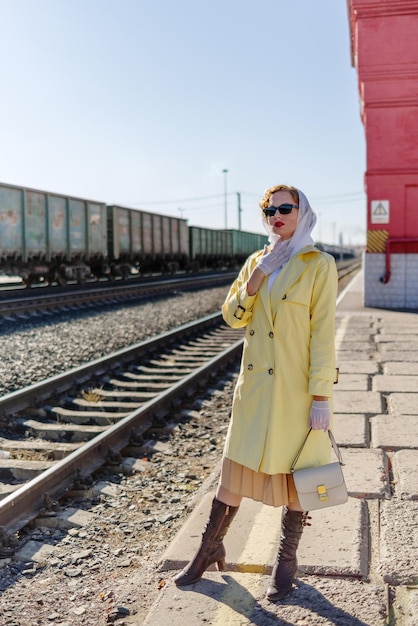 사진 노란색 비옷과 검은 안경을 쓴 젊은 여성이 기차역 플랫폼에 서 있다