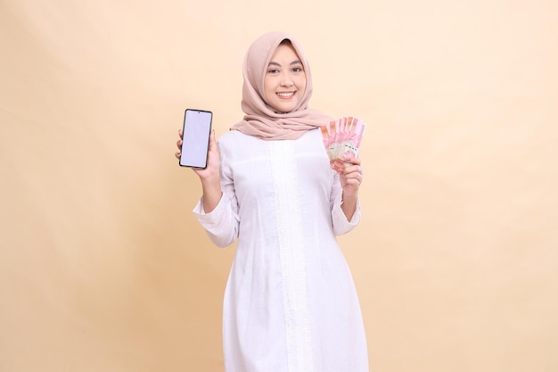 Young woman indonesia muslim wearing a hijab smiles cheerfully holding a cell phone gadget and rupia