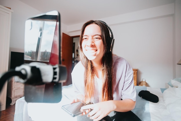 Young woman infuencer talking to followers in front of smart phone while using headphones creating social media content Streaming from home young people new jobs Using phone with tripod