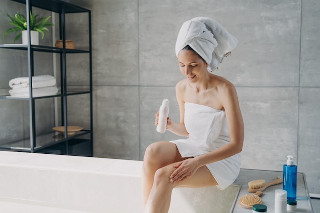 Young woman is bathing in morning at home and applying body lotion to her leg after shower