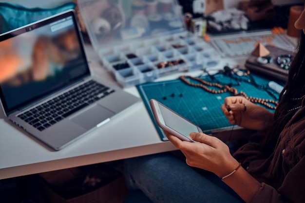 Young woman is chatting by mobile phone while sitting next to table with her jewellery hobby.