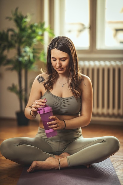 Young woman is doing yoga meditation during coronavirus pandemic in the living room at home. She is ready to take a vitamin drink on floor mat in morning sunshine.