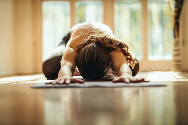 Young woman is doing yoga meditation in the living room at home. She is meditating on floor mat in morning sunshine.