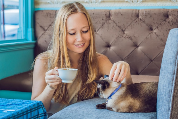 La giovane donna sta bevendo caffè e accarezzando il gatto sullo sfondo del divano graffiato dai gatti