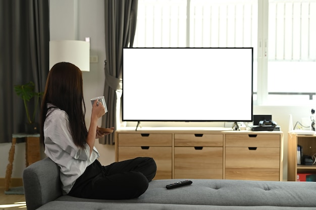 Молодая женщина пьет кофе и смотрит телевизор на диване у себя дома.
