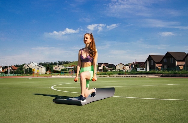 若い女性がサッカー場でフィットネスに従事しています。青い空と明るい太陽、健康的なライフスタイル。