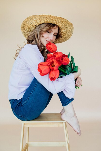 Foto una giovane donna tiene in mano un mazzo di tulipani rossi ed è seduta su una sedia di legno il concetto dell'8 marzo san valentino un regalo meraviglioso per una ragazza ritratto primaverile di una donna con un cappello di paglia