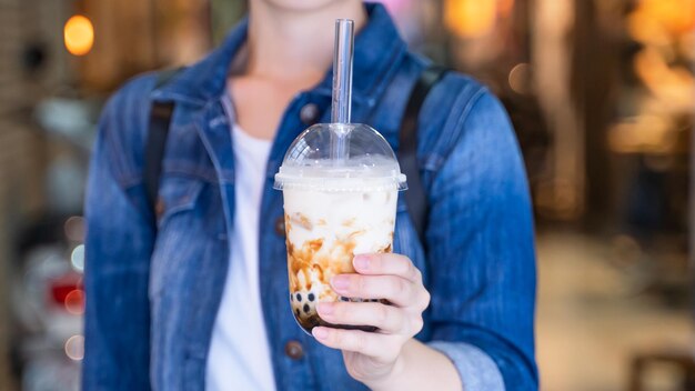 Photo young woman is holding drinking brown sugar flavored tapioca pearl bubble milk tea with glass straw in night market of taiwan close up bokeh