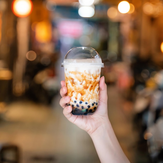 Young woman is holding drinking brown sugar flavored tapioca pearl bubble milk tea with glass straw in night market of Taiwan close up bokeh