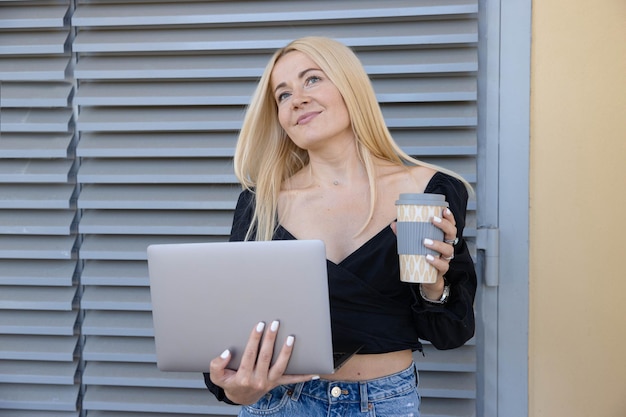 A young woman is holding a laptop and a coffee cup while working outside