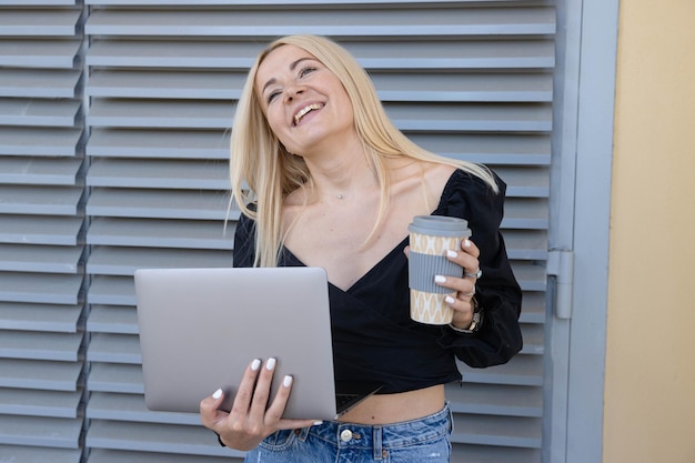 A young woman is holding a laptop and a coffee cup while working outside