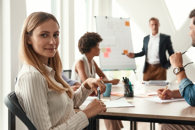 Young woman is looking at camera and smiling while her boss standing near whiteboard and discussing something with team. Teamwork. Meeting