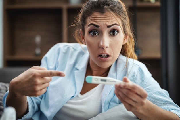 Young woman is lying sick at home couch and looking at thermometer.