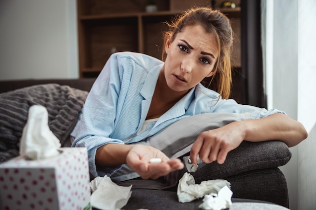 若い女性は自宅のソファで病気で横たわっていて、薬を飲んでいます。