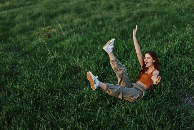 Молодая женщина играет с нами в парке в траве и падает на землю, счастливо улыбаясь на солнечном свете, отдыхая в природе в гармонии с телом.