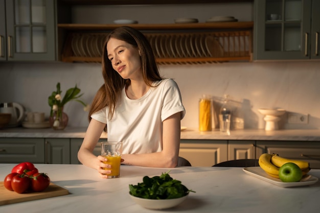 A young woman is seated at a kitchen counter sipping from a glass of fresh juice with a soft morning