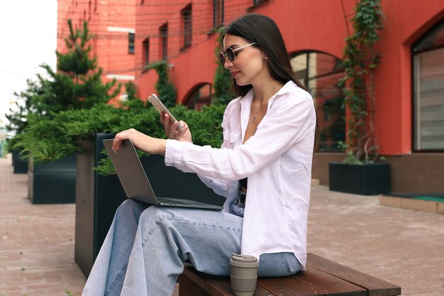 A young woman is sitting on a bench working with a laptop and talking on the phone on the street