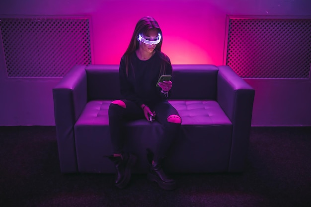 A young woman is sitting on the couch wearing neon glasses and using a gadget high quality photo