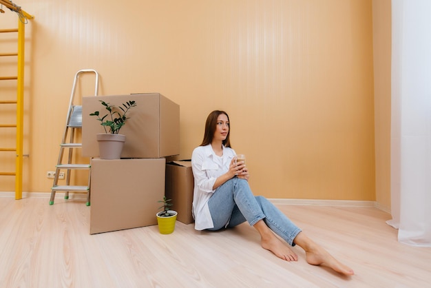 Una giovane donna è seduta per terra davanti agli scatoloni e beve caffè, gioendo e godendosi il nuovo appartamento dopo il trasloco. inaugurazione della casa, consegna e trasporto merci.