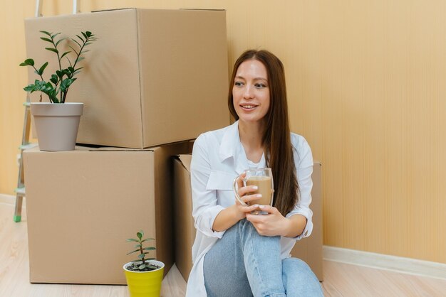 A young woman is sitting on the floor in front of the boxes and drinking coffee, rejoicing and enjoying the new apartment after moving. Housewarming, delivery and cargo transportation.