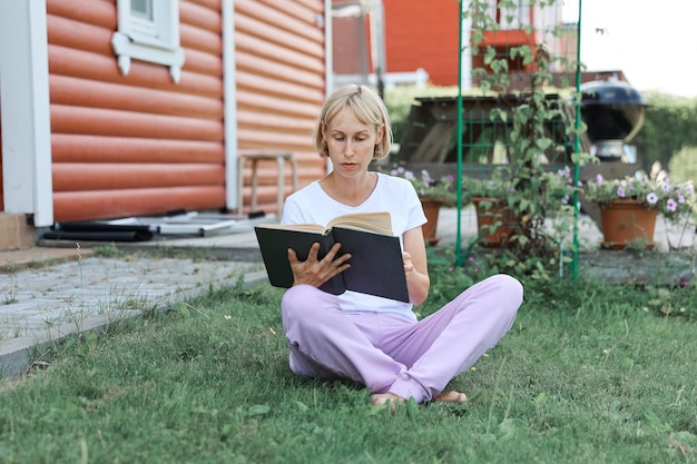 A young woman is sitting on the grass in home yard on a sunny day and reading a real book. Grass and dandilion background, nice vintage look.