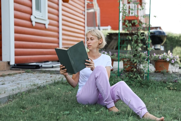 A young woman is sitting on the grass in home yard on a sunny day and reading a real book. Grass and dandilion background, nice vintage look.