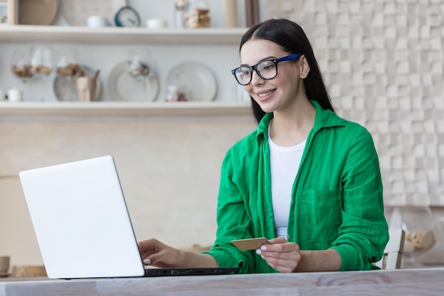 A young woman is sitting in the kitchen at home at the table with a laptop holds a credit card and