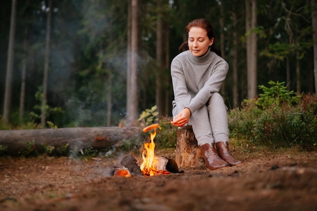 Young woman is sitting on a log in a summer forest cooking food
on a campfire camping in the wild