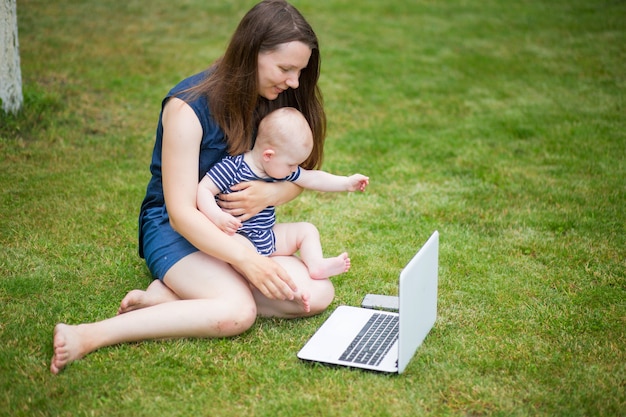 A young woman is sitting in a meadow with a laptop and talking on an online conference with colleagues at work. Remote work in nature. Female student studying at laptop on the grass