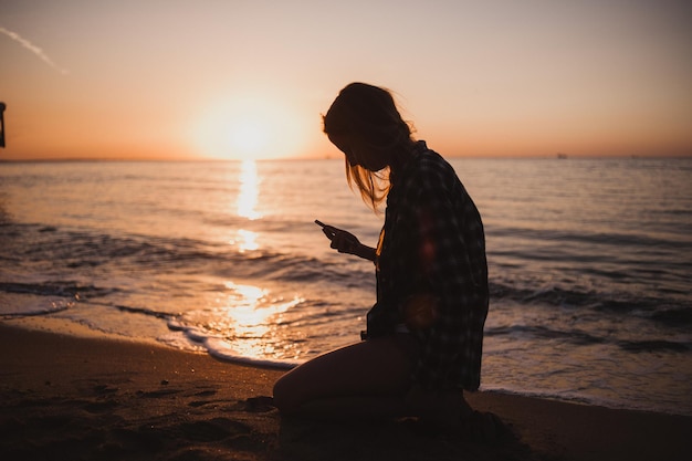 젊은 여성이 석양에 휴대폰을 들고 바다 옆 자갈 해변에 앉아 태양을 배경으로 한 사진