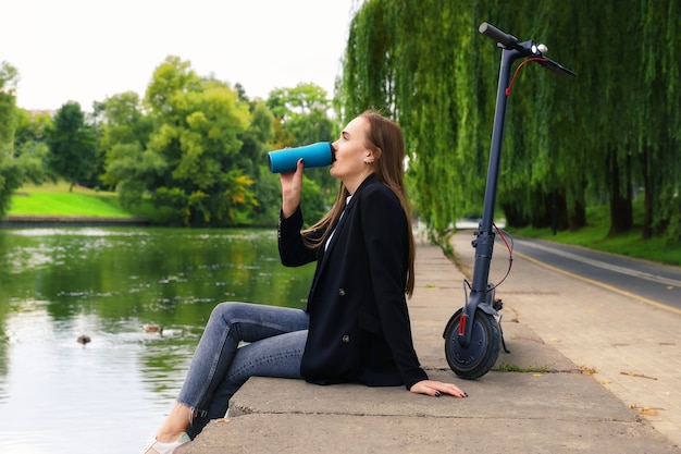 A young woman is sitting on the river embankment and drinking coffee after a trip on an electric scooter