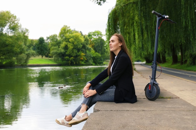 A young woman is sitting on the river embankment next to an electric scooter