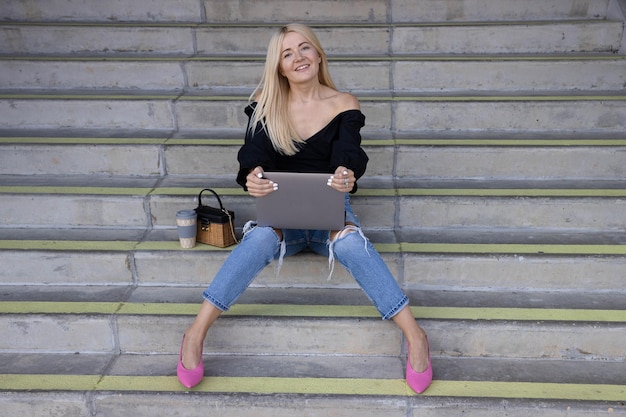 A young woman is sitting on the steps and working on a laptop outside