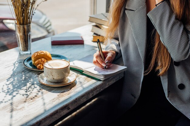 A young woman is sitting at a table by the window and writing in a notebook.