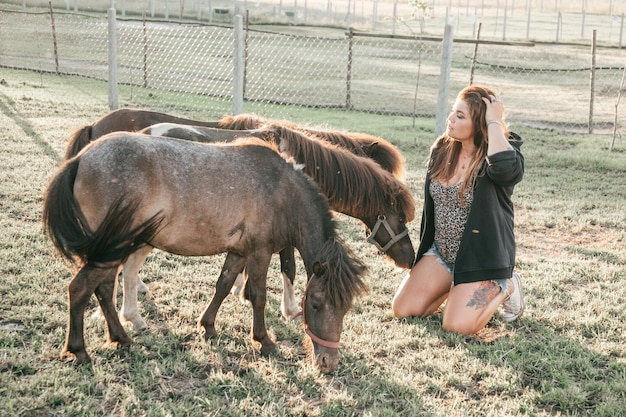 Young woman is sitting next to three grazing ponies . life on\
farm. agrotourism. beautiful animals . summer holidays. own farm.\
shaggy head.funny animals. hippotherapy.