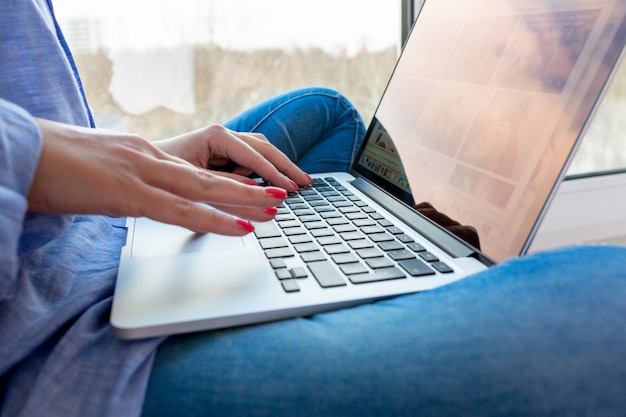 Young woman is sitting on the windowsill and working at laptop