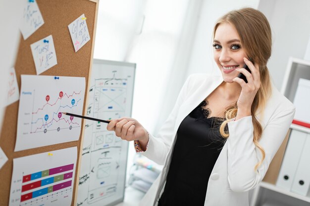 A young woman is standing near the board with stickers, talking on the phone and holding a pencil in her hand.