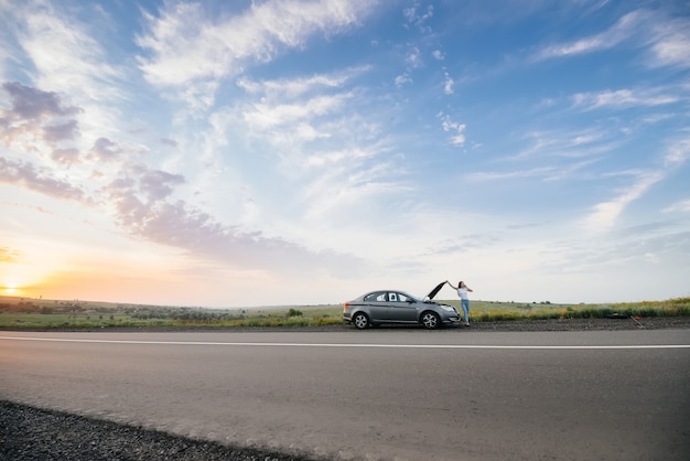 A young woman is standing near a broken-down car.

