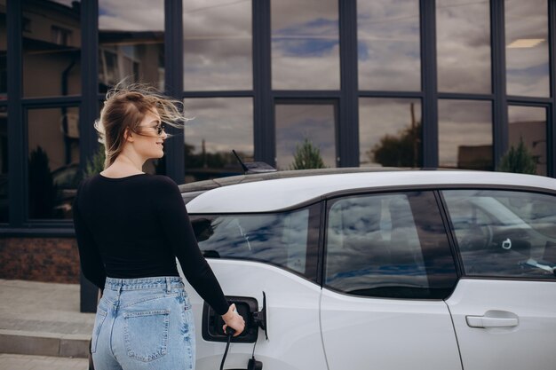 Young woman is standing near the electric car and looks at the\
smart phone the rental car is charging at the charging station for\
electric vehicles car sharing