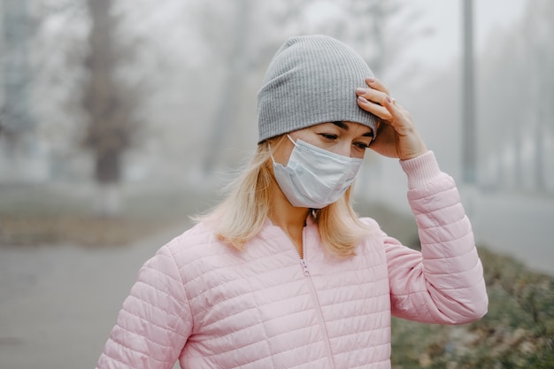 若い女性が彼女の手で彼女の頭をつかんで医療マスクで道路の近くに立っています。路上で冬の頭痛。