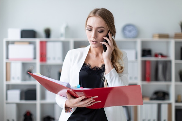 A young woman is standing in the office next to the shelving, talking on the phone and holding a folder with documents.