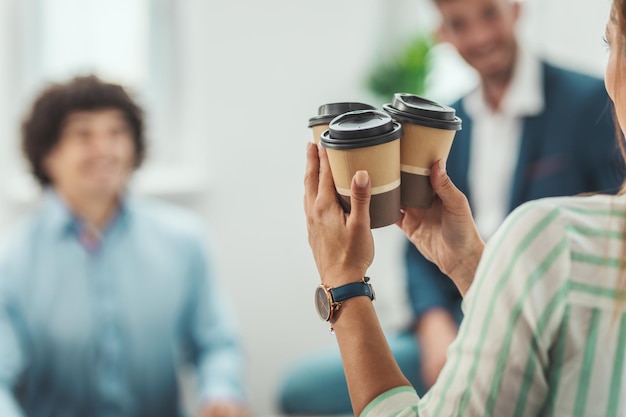 A young woman is taking disposable coffee cups to her smiling colleagues during coffee break in the office.