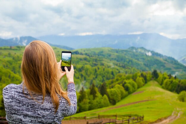 Молодая женщина фотографирует удивительный красивый пейзаж с горным лесом и облачным небом