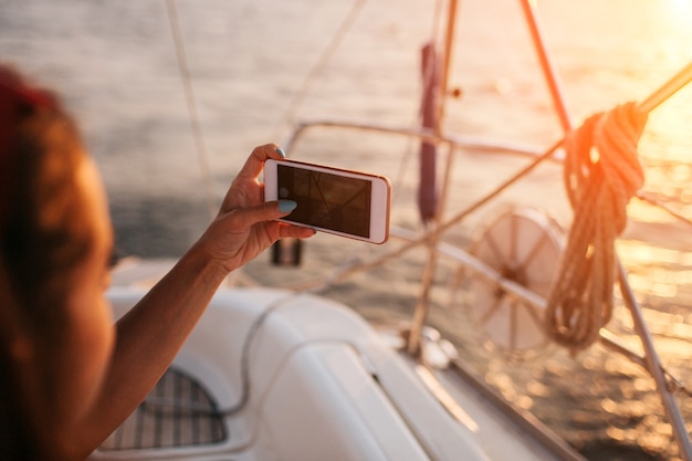 Young woman is taking picture of beautiful and orange sunset upon river. She holds phone in hand and look forward. Woman is on yacht