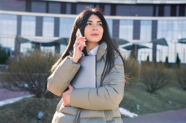 A young woman is talking on the phone and holding a laptop outside