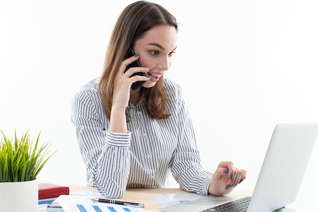 A young woman is talking on the phone and typing on a laptop while sitting at her workplace Business portraits in the studio on a white background